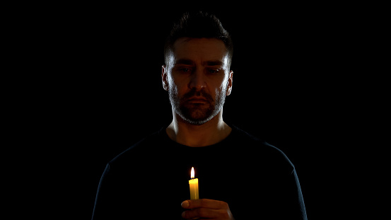 Desperate man holding candle on black background, symbol of hope, sorrow ritual