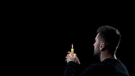 Praying man holding burning candle on dark background, spiritual ritual, faith