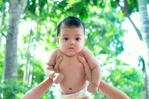 o novo nascido do sudeste asiático não está vestindo camisa. o recém-nascido do sudeste asiático é 3 meses velho. criança da terra arrendada da matriz com mãos. o lado traseiro do miúdo novo é árvores. o fundo da criança é verde & frescor. - southeast asian ethnicity fotos - fotografias e filmes do acervo