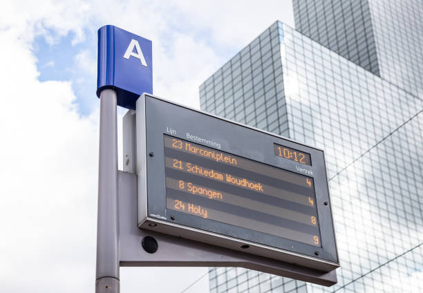 Rotterdam, Netherlands - July 1, 2019: Low angle of electronic information board at public transport stop beside skyscraper over cloudy sky