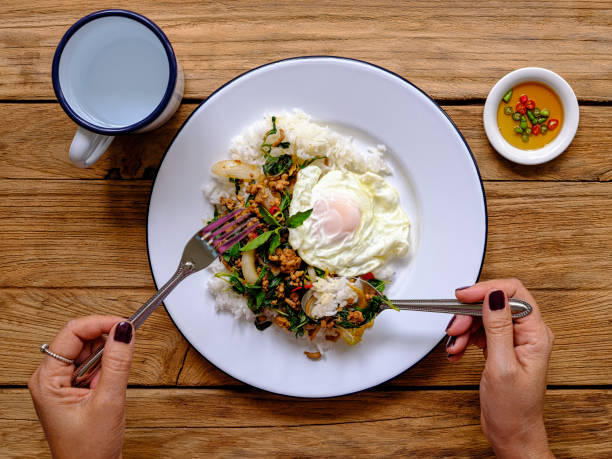 This delicious dish is made of fresh minced pork, cooked in hot oil in a wok mixed with fresh holy basil, fresh chili, onion and garlic with seasoning. This dish is normally served on top of fresh steamed rice with a fried egg and chili with fish sauce as complements to the dish. This popular Thai recipe is found across Thailand being recognized as a quick tasty dish, it can be ordered in restaurants but is well known for being a popular street food dish. This dish is photographed with a spoon and a fork being used to eat the food with, this is traditionally what is used in Thailand to eat Thai food with, the spoon replaces the knife, as used in many western countries. el famoso plato tailandés picante llamado 'pak krapao moo', cerdo frito con albahaca sagrada, en inglés, servido en una cama de arroz al vapor con un huevo frito y 'pik nam bla' (chili con salsa de pescado) en una vieja mesa de madera, siendo comido por