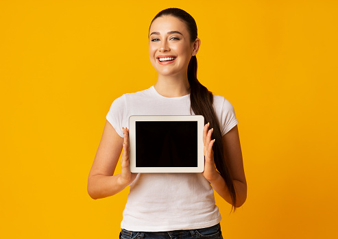 Girl Showing Blank Tablet Screen On Yellow Background. Studio Shot, Free Space, Mockup