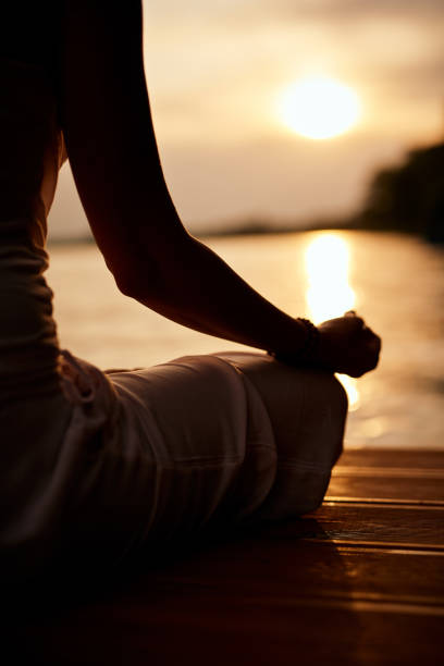 Rear view of Caucasian woman sitting in lotus position and meditating on dock at sunset.