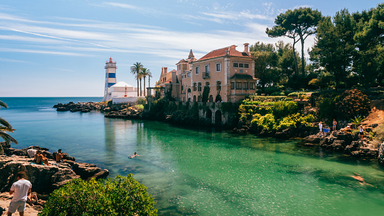 Cascais, Lisbon, Portugal - August 29, 2019: View of Santa Marta lighthouse and Municipal museum of Cascais, in Portugal with tourists bathing in the beautiful green water on a sunny summer day