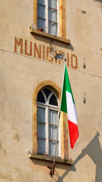 Close-up view of an Italian flag placed on a window sill with red and white flowers.The facade of the old building is painted ochre-coloured. Square composition and copy space.