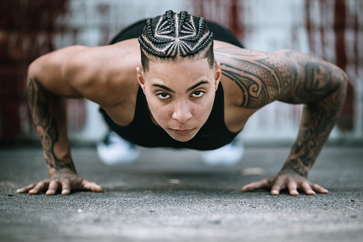 An mixed race adult woman with muscular build poses for a portrait in a grunge urban setting.  She wears athletic clothing, sporting a unique braid pattern in her hair and a sleeve tattoo.