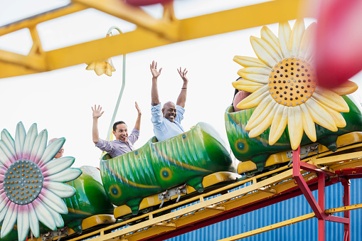 Two mature men at an amusement park participating in corporate team building activities.  They are sitting on a small rollercoaster, smiling with their arms raised over their heads. The African-American man who is looking at the camera is in his 50s. His coworker is Hispanic, in his 40s.