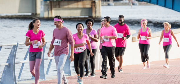 Father and daughter walking in breast cancer race A mature man in his 40s walking beside his mixed-race African-American and Caucasian 13 year old teenage daughter. They are leading a group of nine men and women wearing pink shirts, participating in a charity event to raise money for breast cancer research. They are in a race, wearing numbered marathon bibs. power walking photos stock pictures, royalty-free photos & images