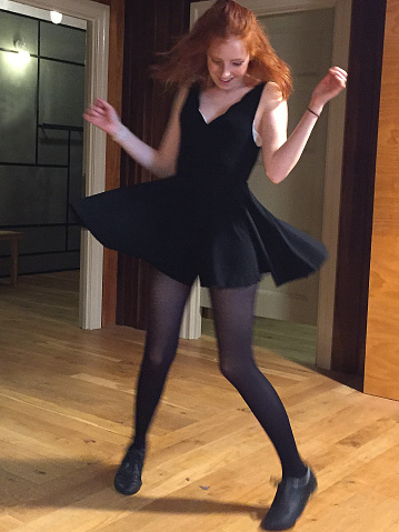 Stock photo of young teenage redhead girl spinning around in little black dress with shoulder straps and scalloped V-neck / LBD party dress, teenager dancing and looking happy with red ginger hair, freckles and natural makeup, black tights on wooden dance floor
