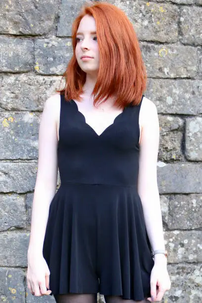 Photo of Image of red haired teenage girl 14 / 15 with pale skin and freckles stood outdoors before an old stone wall covered in lichen, wearing a smart casual black playsuit with scalloped v-neckline and modelling a choppy bob hairstyle