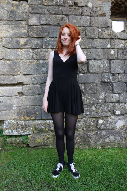 Stock photo showing a full-length image of a beautiful, ginger haired teenager modelling a choppy bob hairstyle with pale skin and freckles stood outdoors before an old stone wall. The model is wearing a smart casual black playsuit with scalloped v-neckline, black tights and black pumps with white laces. imagen de cuerpo entero de adolescente de pelo rojo 14 / 15 con piel pálida y pecas estaba al aire libre antes de una vieja pared de piedra cubierta de líquenes, usando un elegante traje de juego negro casual con escote en v escaldado y modelando un pein