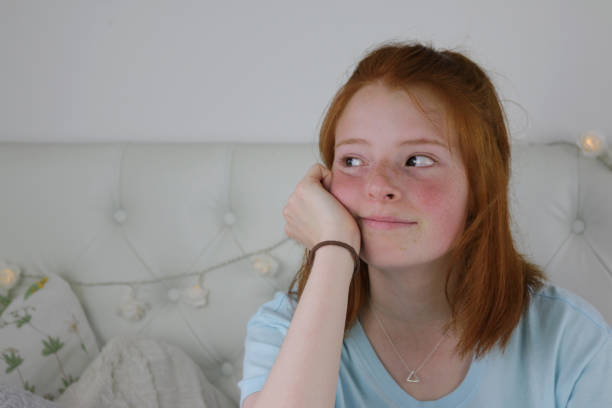 Image of red haired teenage girl 14 / 15 with pale skin, freckles and flushed cheeks sat in bedroom looking bored and fed up, chin leaning on hand looking glum Stock photo showing a ginger haired teenager in her bedroom head on hand looking fed up and frustrated. rosy cheeks stock pictures, royalty-free photos & images