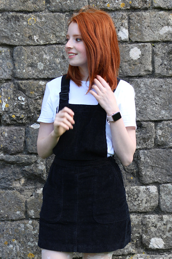 Stock photo showing a full-length image of a beautiful, ginger haired teenager modelling a choppy bob hairstyle with pale skin and freckles stood outdoors before an old stone wall. The model is wearing a smart casual black, ribbed corduroy pinafore dress over a white t-shirt, white patterned tights and black suede shoes.