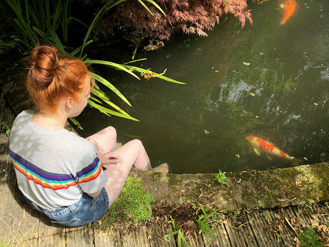 Stock photo showing high-angle view of ginger haired teenager sitting on the edge of a fish pond dangling her feet in the water.