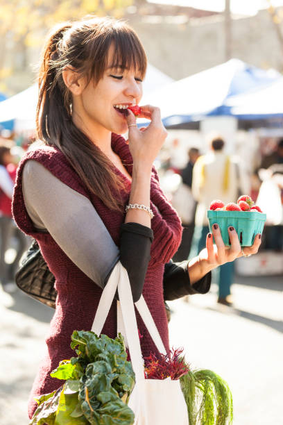 beautiful, happy, smiling, young hispanic woman eating fresh, organic strawberries fruit at farmers market. healthy lifestyles. - market fruit strawberry farmers market imagens e fotografias de stock