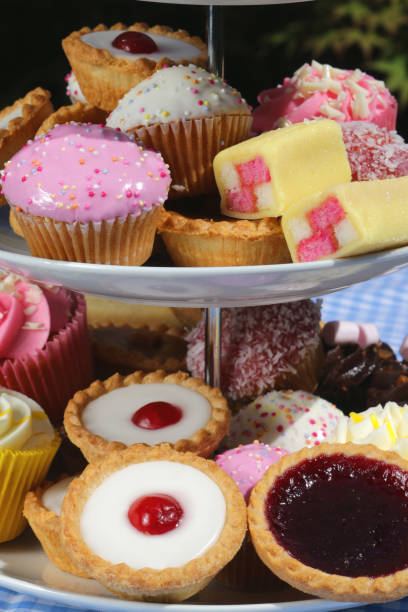 imagem do piquenique do bufete do chá da tarde dos bolos dos queques no carrinho estratificado do bolo da porcelana branca com três camadas no tablecloth checkered azul no partido do jardim, as tortas do bakewell da cereja, muffins do chocolate, bolo cas - fondant fancies - fotografias e filmes do acervo