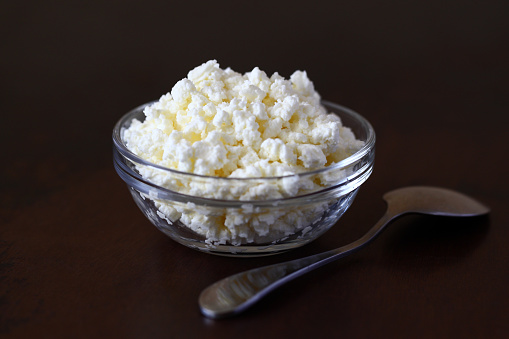 Cottage cheese crumbly in a glass transparent bowl and metal spoon on a wooden old scratched brown tabletop.