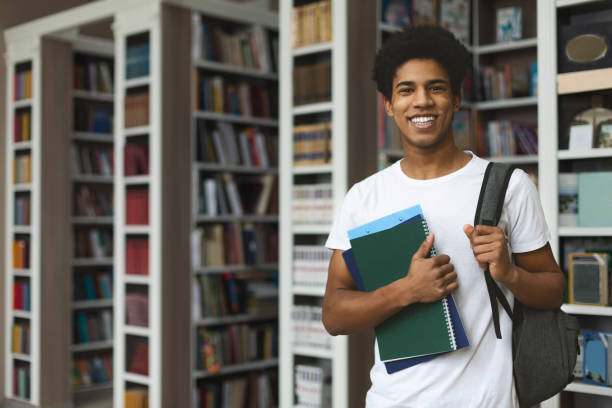 Handsome afro student posing on bookshelves background Handsome african american male student posing in campus library, empty space textbook photos stock pictures, royalty-free photos & images