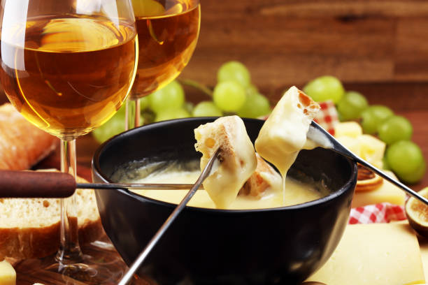 dîner de fondue suisse gastronomique un soir d'hiver avec des fromages assortis sur une planche à côté d'une marmite chauffée de fondue au fromage avec trempette à deux fourchettes - fondue fork photos et images de collection