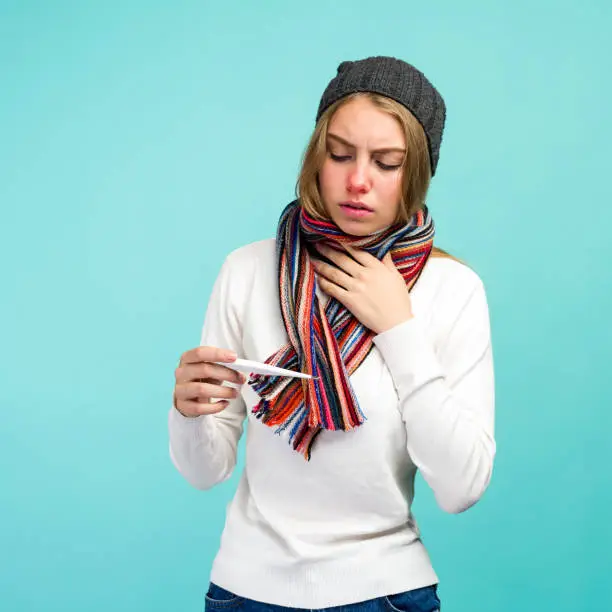 Sad teen girl having flue taking thermometer against blue background. Beautiful lady is sick with a high temperature, isolated close-up. - image