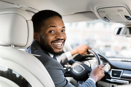 Young afro businessman at driver's seat smiling to camera over shoulder, free space
