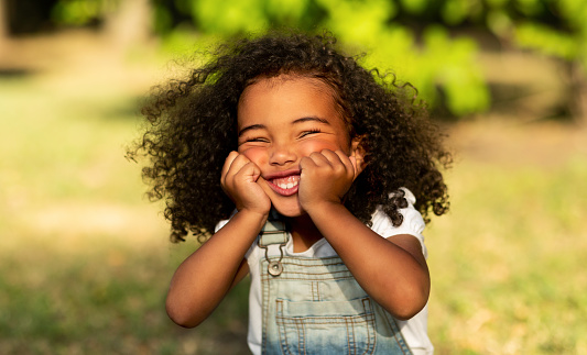 Funny little afro girl touching cheeks and having fun in nature park