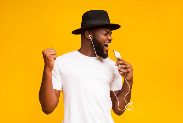 Happy african man in hat singing into smartphone like microphone Emotional african guy in black hat singing into smartphone like microphone and listening to music via earphones, yellow background with free space happy people audio stock pictures, royalty-free photos & images