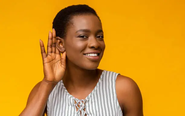 Curious interested african american woman holding hand near ear and trying to hear information, yellow studio background