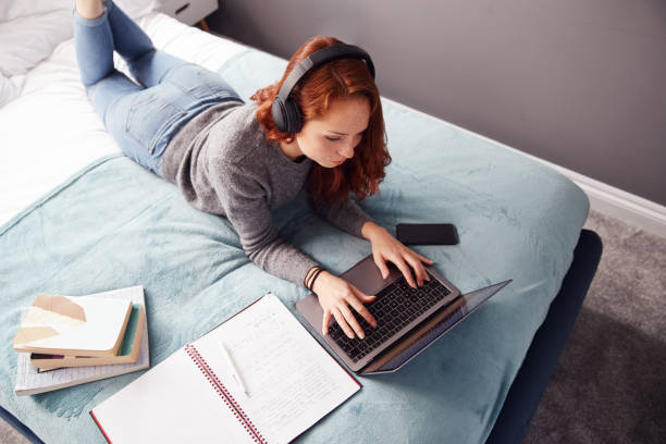 Looking Down On Female College Student Wearing Headphones Lying On Bed Working On Laptop Looking Down On Female College Student Wearing Headphones Lying On Bed Working On Laptop britain british audio stock pictures, royalty-free photos & images