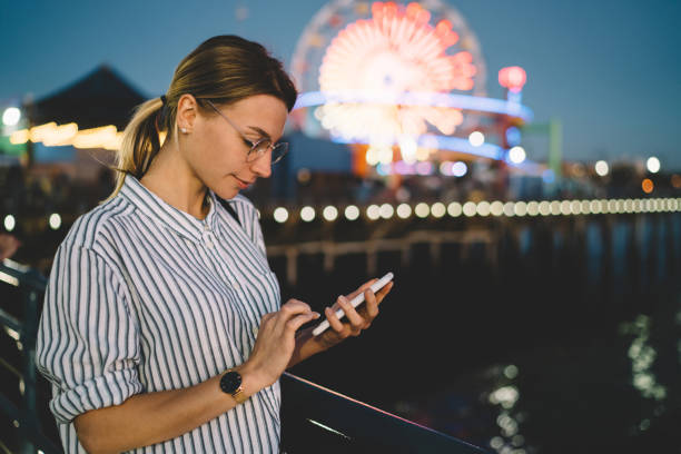 young female blogger share content in networks via smartphone recreating in santa monica pier in evening,hipster girl texting message on cellphone standing on beach amusement park shining lights - santa monica pier santa monica beach night amusement park imagens e fotografias de stock