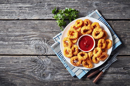 crispy calamari rings, deep-fried breaded squid rings served with tomato sauce on a white plate on an old wooden table, horizontal view from above, flat lay, free space