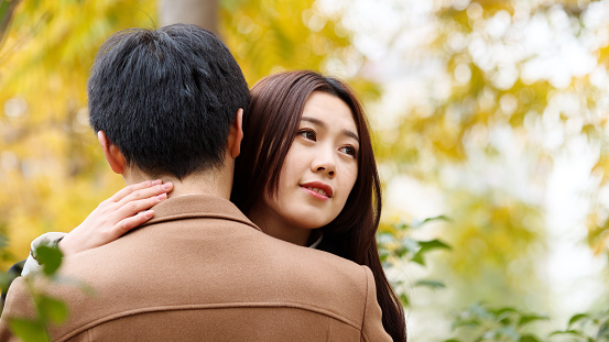 Beautiful Chinese young woman hugging her boyfriend's neck and looking away full of happiness with autumn yellow background, lover concept.