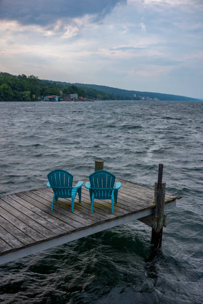 Rough waters, crash into a pier at Seneca Lake at Watkins Glen in New York State. Rough waters, crash into a pier with two plastic and blue Adirondack garden chairs, as a storm comes in over Seneca Lake at Watkins Glen in New York State. finger lakes stock pictures, royalty-free photos & images