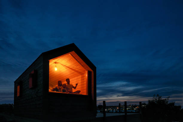 una famiglia di tre persone è seduta su un letto vicino alla finestra, leggendo un libro e parlando. vista dell'esterno della casa. cielo blu notturno. luce calda arancione da una lampada a incandescenza. - outdoors looking at camera little girls child foto e immagini stock