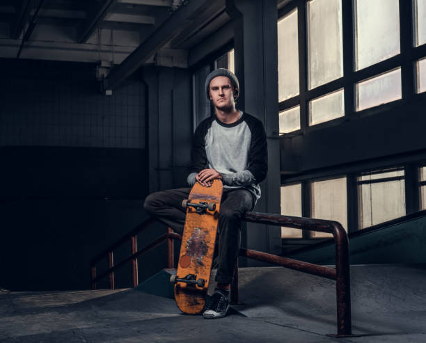 Young handsome man holding his board while sitting on a grind rail in skatepark indoors