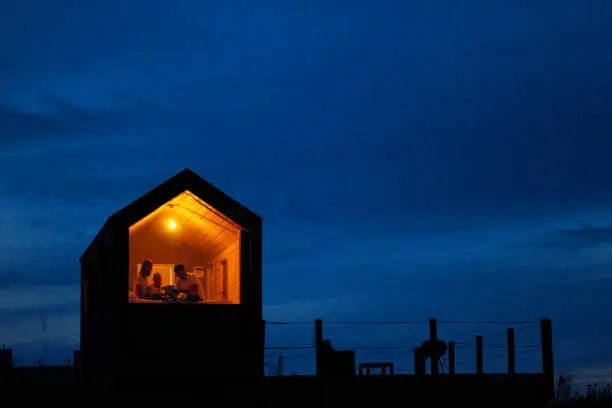 A family of three is sitting on a bed by the window, reading a book and talking. View of the outside of the house. Night blue sky. Orange warm light from an incandescent lamp. The concept of home comfort.