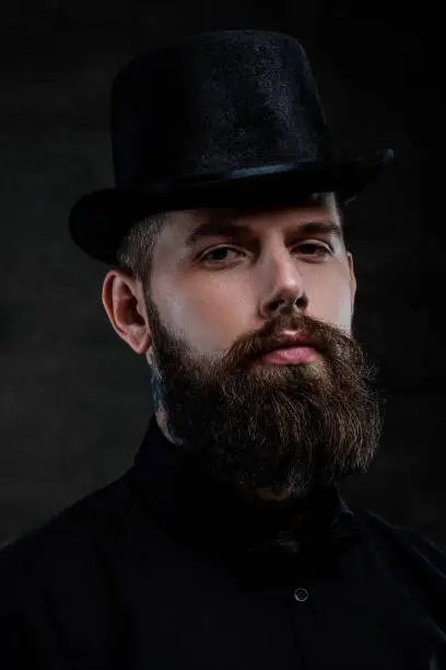 Photo of Close-up portrait of an old-fashioned bearded man with tattoos on his neck wearing a top hat, isolated on a dark background. Studio shot