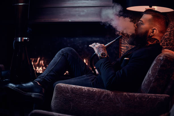 Bearded relaxed man is sitting on the armchair and smoking hookah near fireplace stock photo