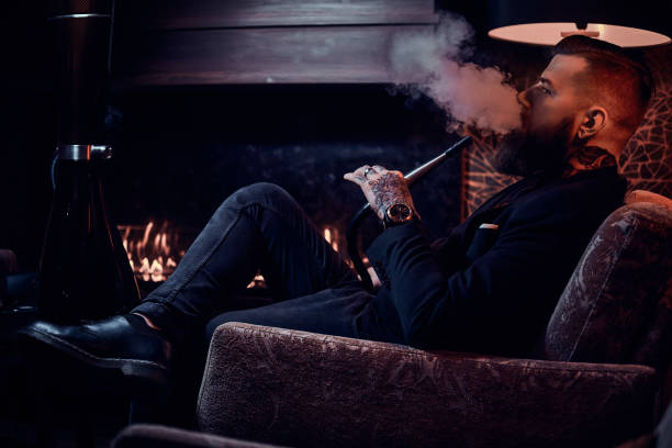 Bearded relaxed man is sitting on the armchair and smoking hookah near fireplace Bearded relaxed man is sitting on the armchair and smoking hookah near fireplace. hookah smoking smoke men stock pictures, royalty-free photos & images