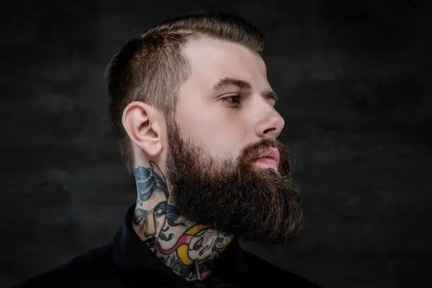 Photo of Profile portrait of an expressive bearded man with tattoos on his neck, isolated on a dark background.