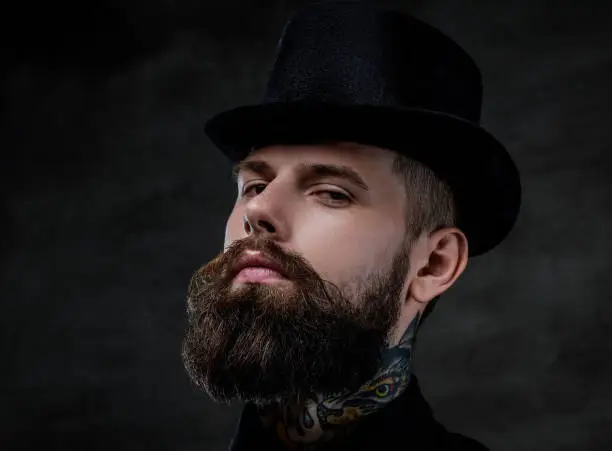 Photo of Close-up portrait of an old-fashioned bearded man with tattoos on his neck wearing a top hat, isolated on a dark background. Studio shot