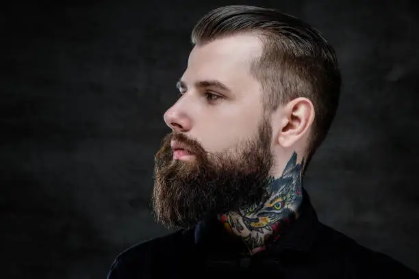 Photo of Profile portrait of an expressive bearded man with tattoos on his neck, isolated on a dark background.