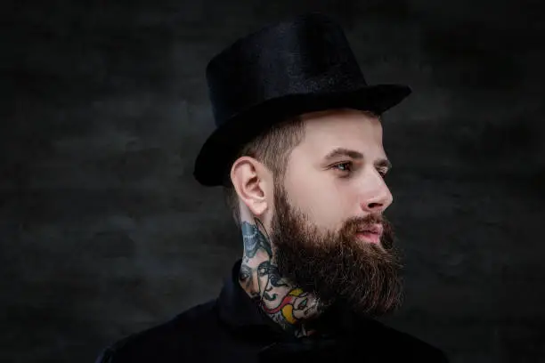 Photo of Close-up portrait of an old-fashioned bearded man with tattoos on his neck wearing a top hat, isolated on a dark background. Studio shot