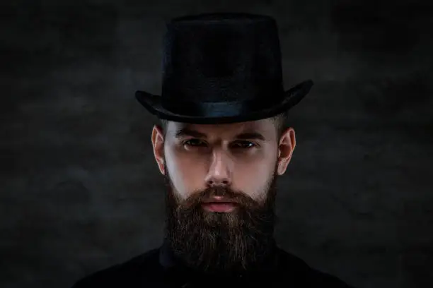 Photo of A serious old-fashioned bearded man wearing a top hat, looking at a camera, isolated on a dark background.