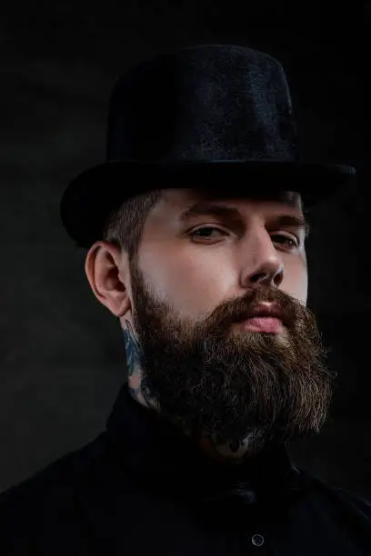 Photo of Close-up portrait of an old-fashioned bearded man with tattoos on his neck wearing a top hat, isolated on a dark background. Studio shot