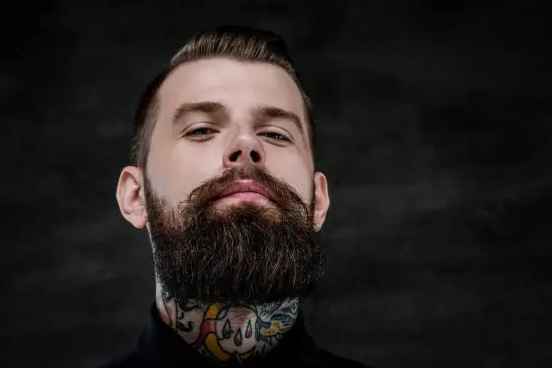 Photo of Close-up portrait of an expressive bearded man with tattoos on his neck, isolated on a dark background. Studio shot