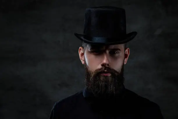 Photo of A serious old-fashioned bearded man wearing a top hat, looking at a camera, isolated on a dark background.