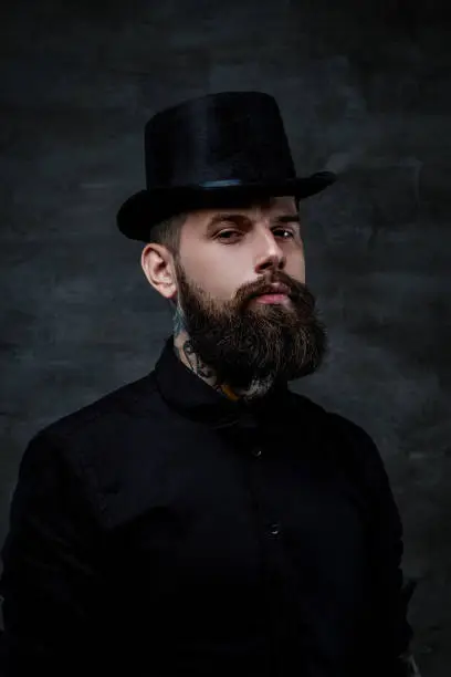 Photo of Portrait of an old-fashioned bearded man with tattoos on his neck wearing a top hat, isolated on a dark background. Studio shot