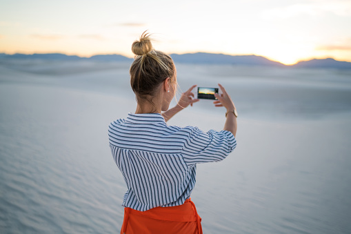 Back view of young woman using smartphone for making selfie standing at beautiful white sand dunes at sunset, hipster girl photographing of scenic landscape on sundown enjoying wanderlust vacations