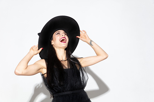 Young brunette woman in black hat and costume on white background. Attractive caucasian female model. Halloween, black friday, cyber monday, sales, autumn concept. Copyspace. Laughts, scary.